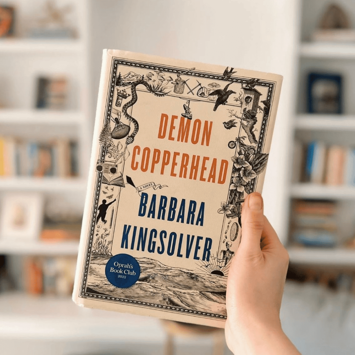 person holding demon copperhead by barbara kingsolver in front of bookshelves.