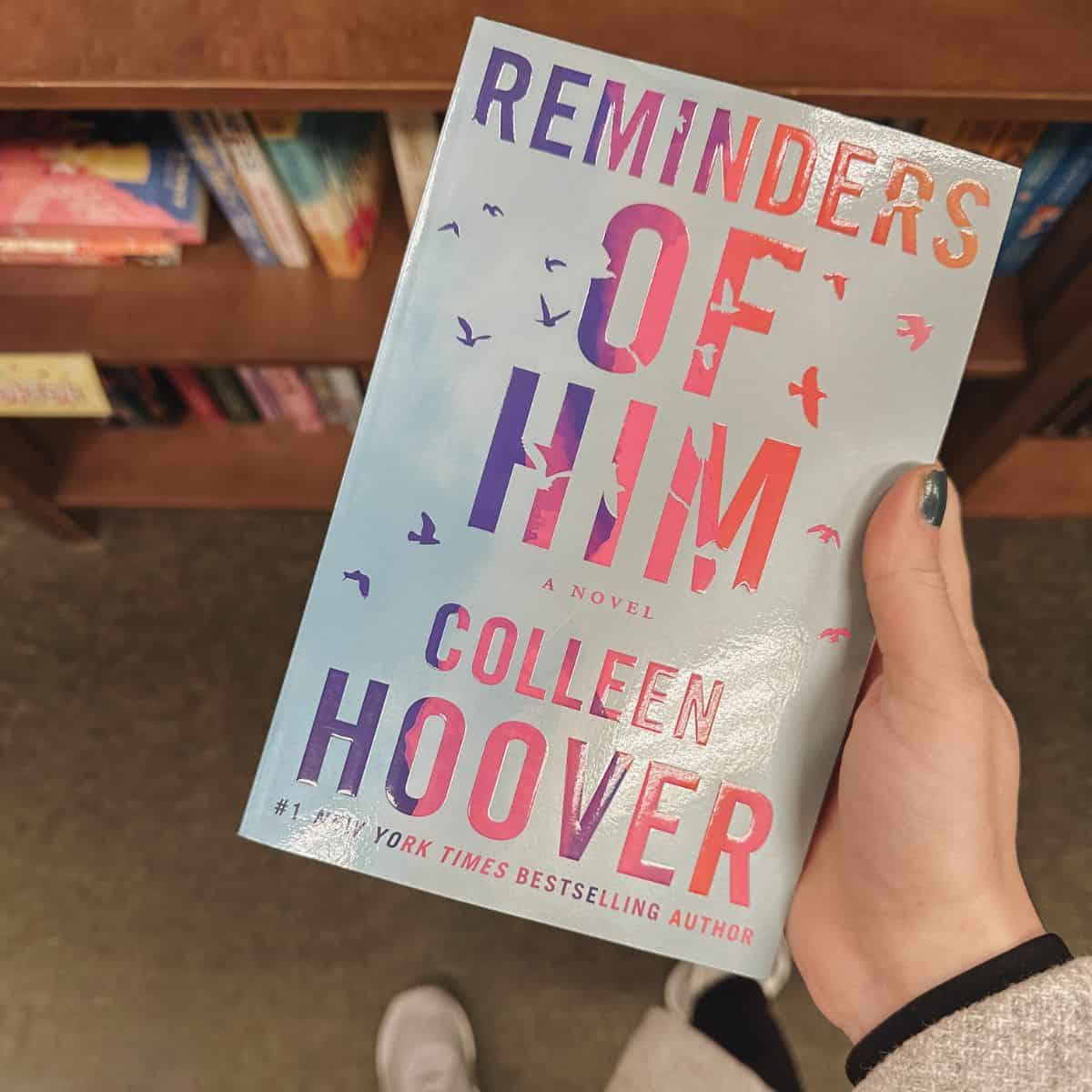 reminders of him by colleen hoover in front of bookshelves.