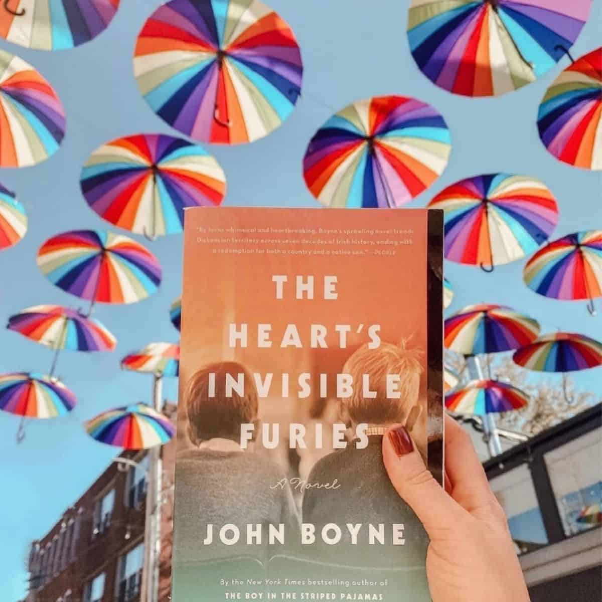 person holding the hearts invisible furies by john boyne in front of rainbow umbrellas.