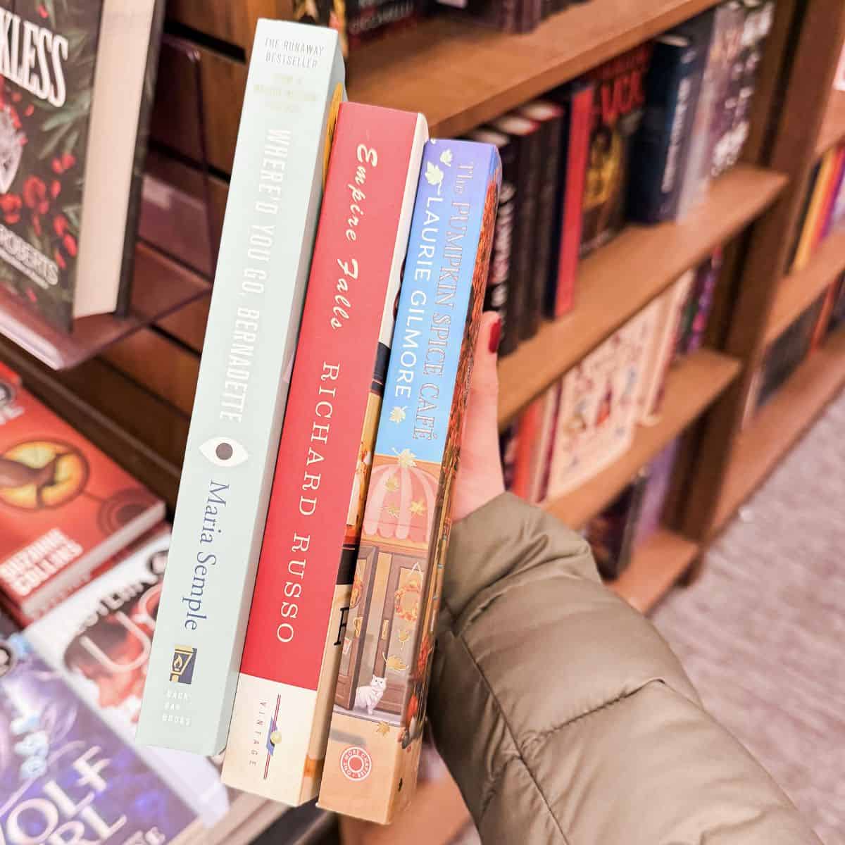 person holding three books like gilmore girls in front of a bookshelf.